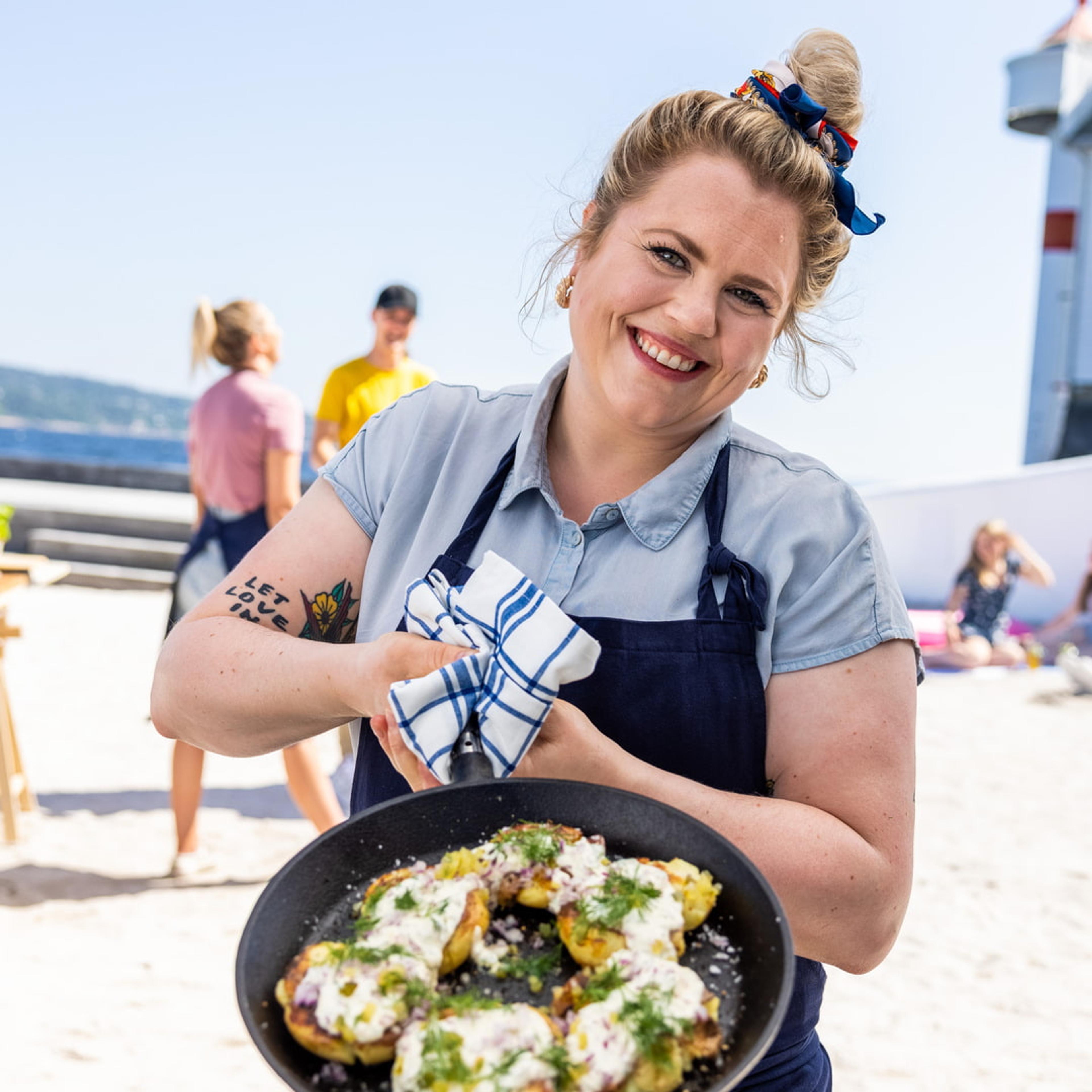 Reneé Fagerhøi holder fram en panne med smasha poteter toppet med rømmedressing, urter og rødløk, på en strand i solskinn