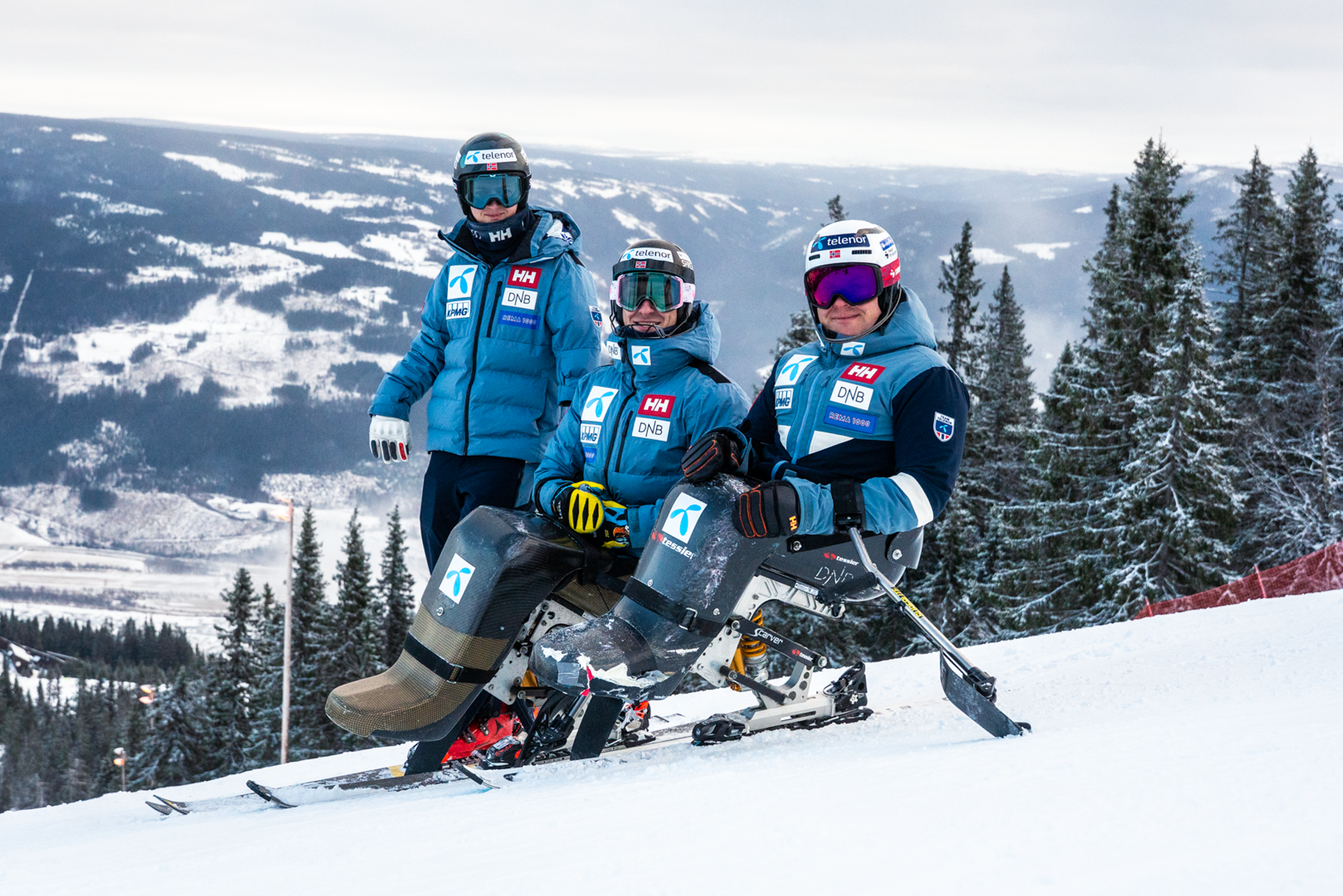 Tre parautøvere i blå landslagsbekledning på en snødekt alpinbakke. To av utøverne sitter i sitski, mens den tredje står ved siden av. Snødekte fjell og grantrær strekker seg utover i bakgrunnen.
