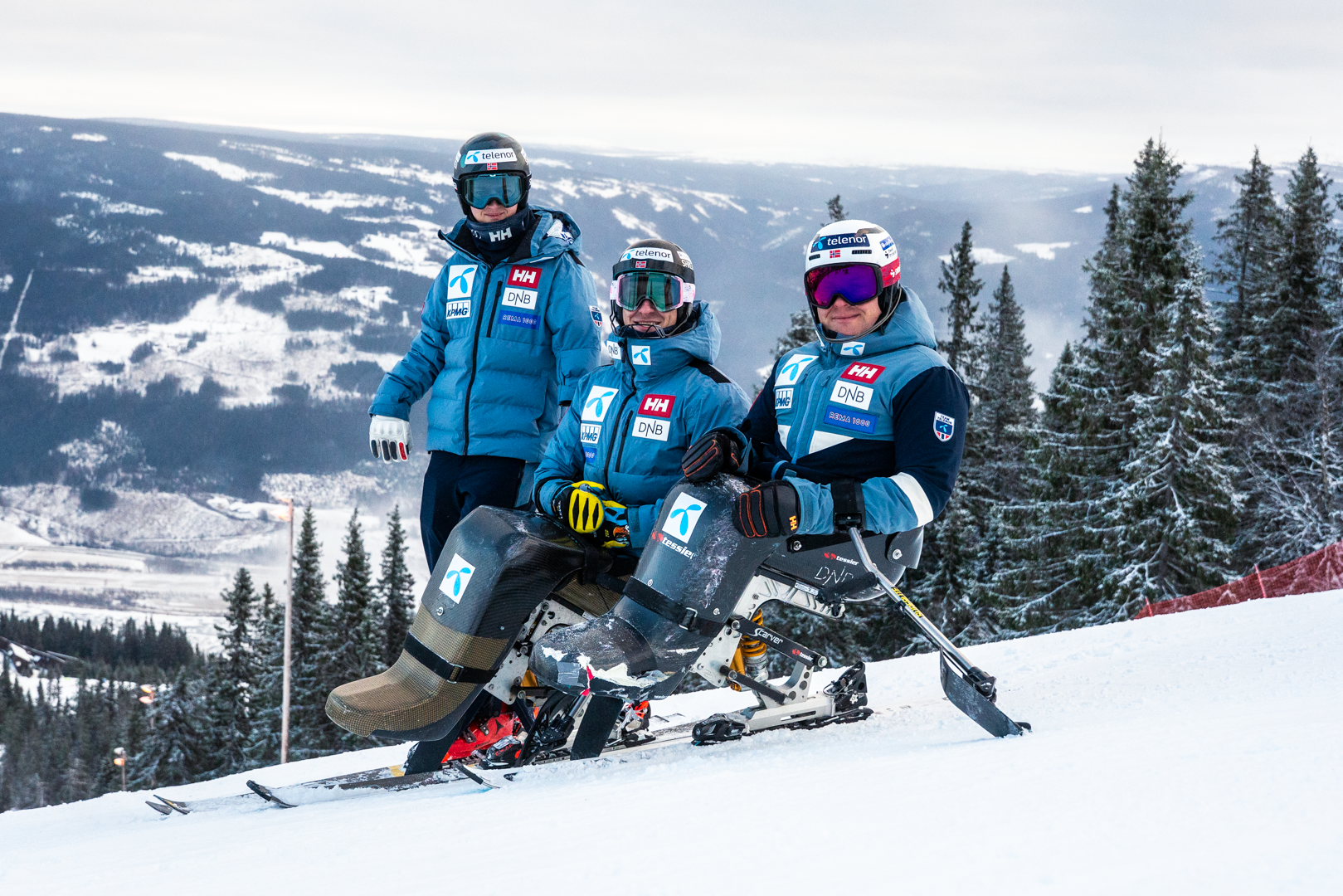 Tre parautøvere i blå landslagsbekledning på en snødekt alpinbakke. To av utøverne sitter i sitski, mens den tredje står ved siden av. Snødekte fjell og grantrær strekker seg utover i bakgrunnen.