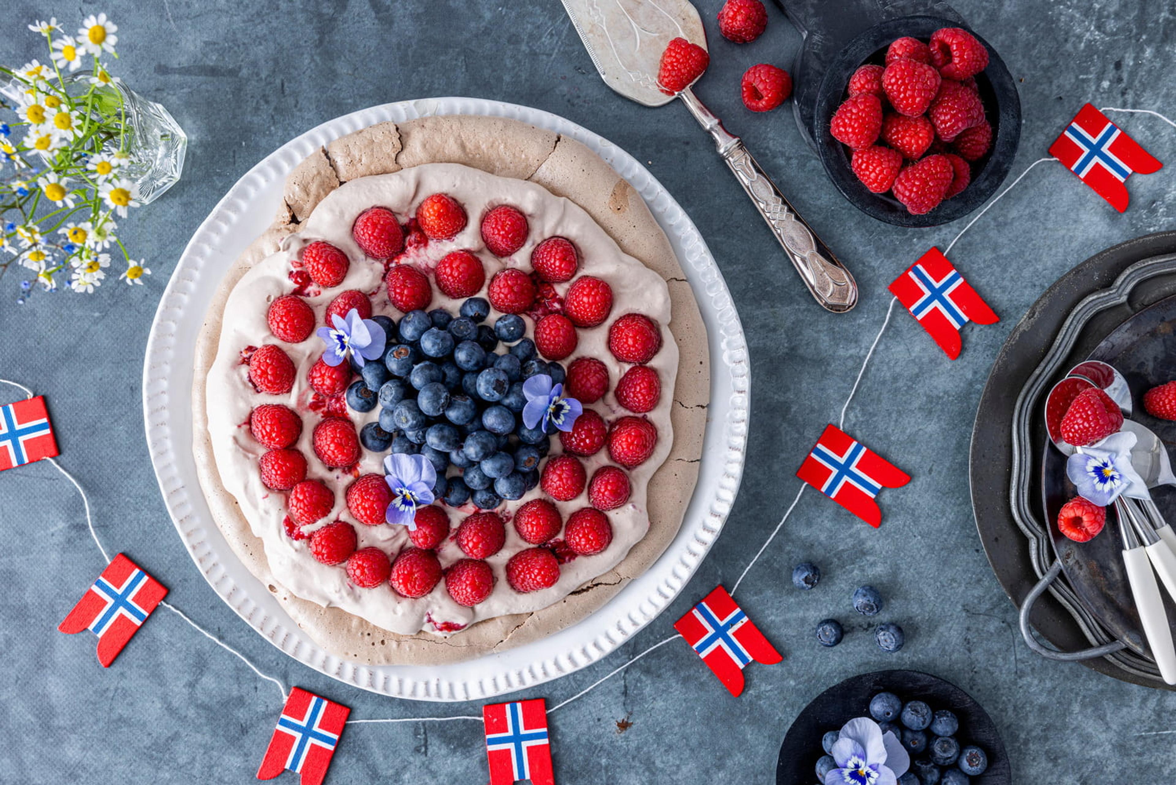 Pavlova pyntet med masse bringebær og blåbær, på et bord pyntet med flagg-girlander i anledning 17. mai