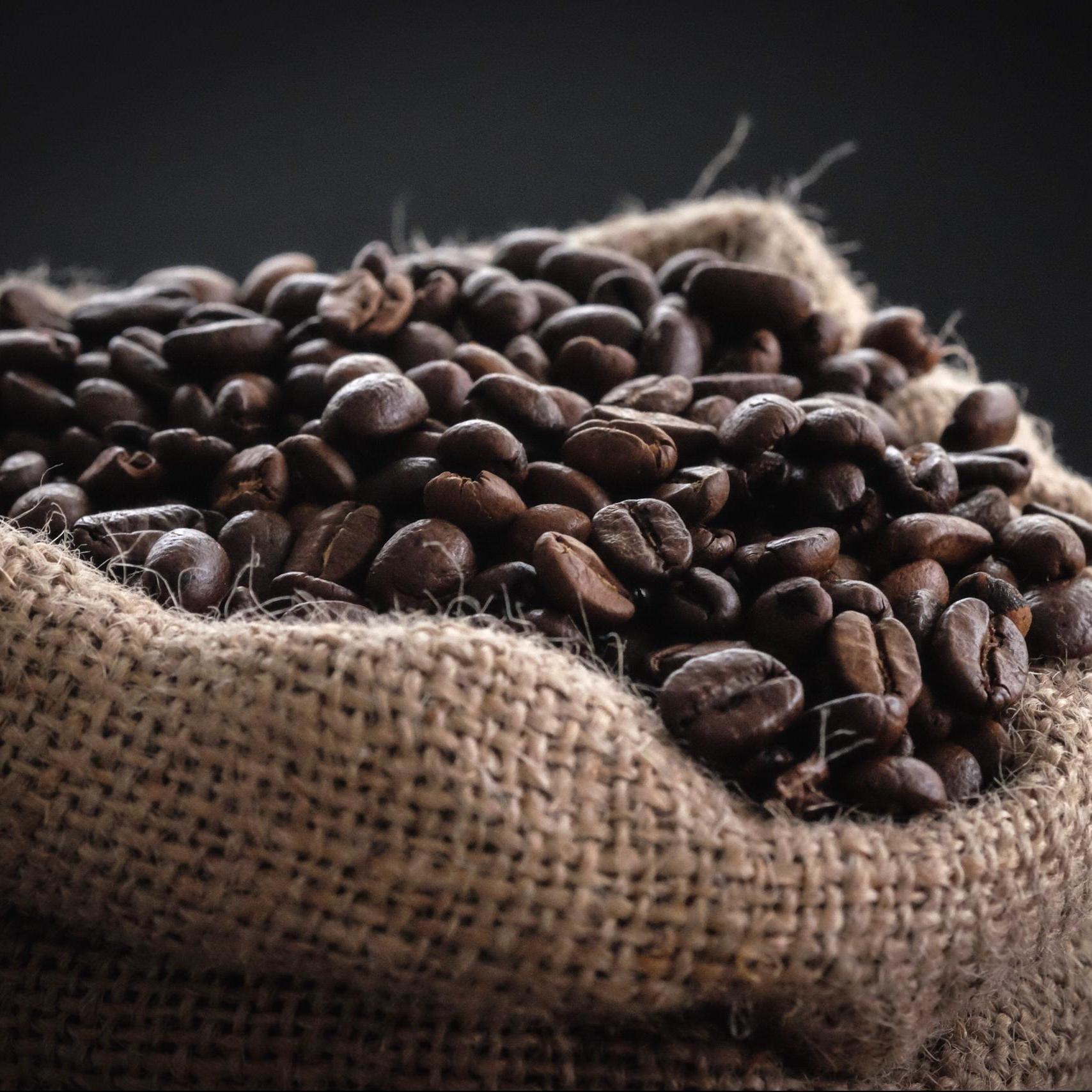 Nærbilde av mørkristede kaffebønner i en sekk av grovt jutestoff. Kaffebønnene fyller sekken og er fotografert mot en mørk bakgrunn som fremhever farge og tekstur.