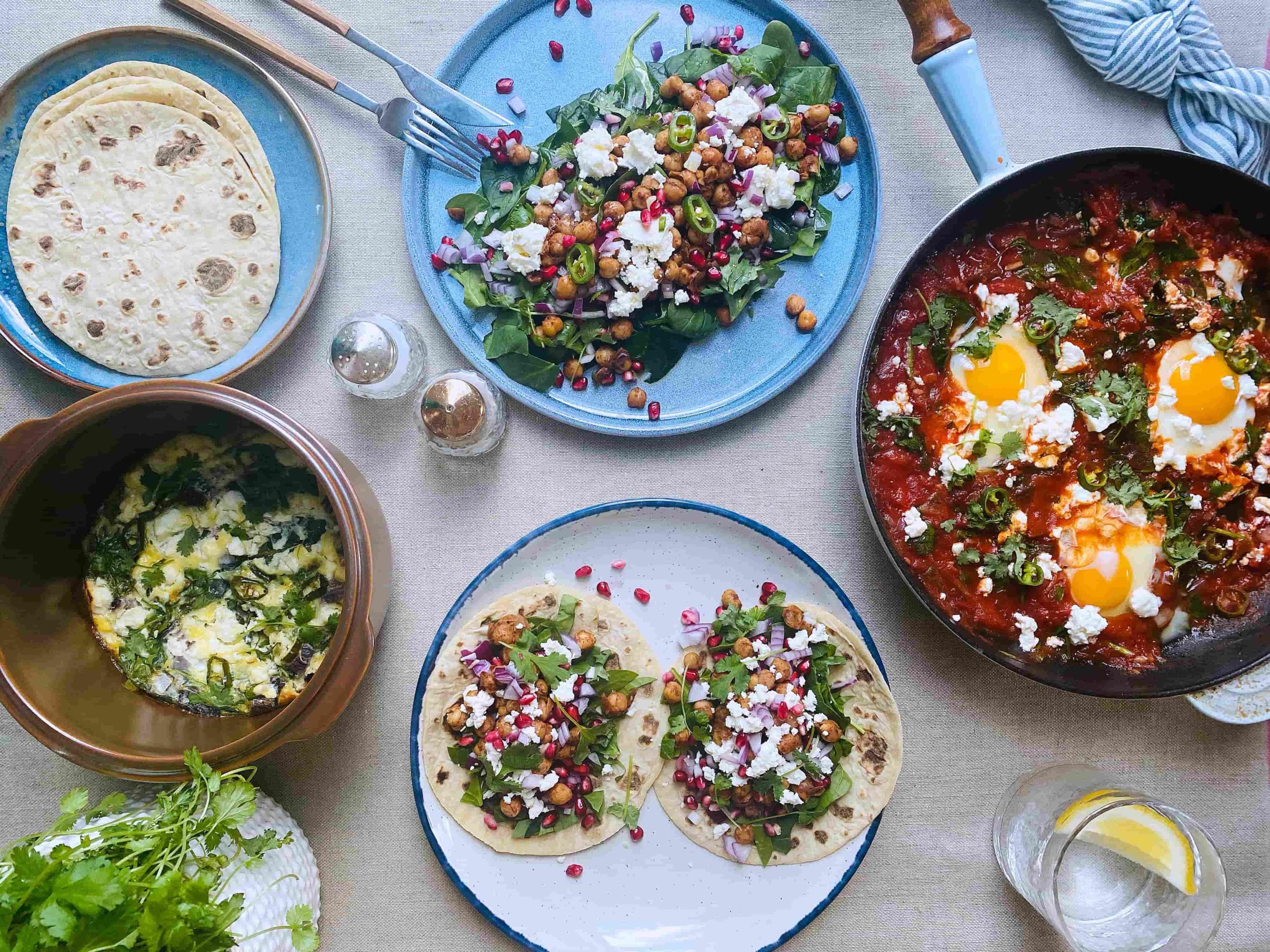 Et dekket bord sett ovenfra med fargerike retter, inkludert shakshuka i en panne, kikertsalat og fylte tortillas