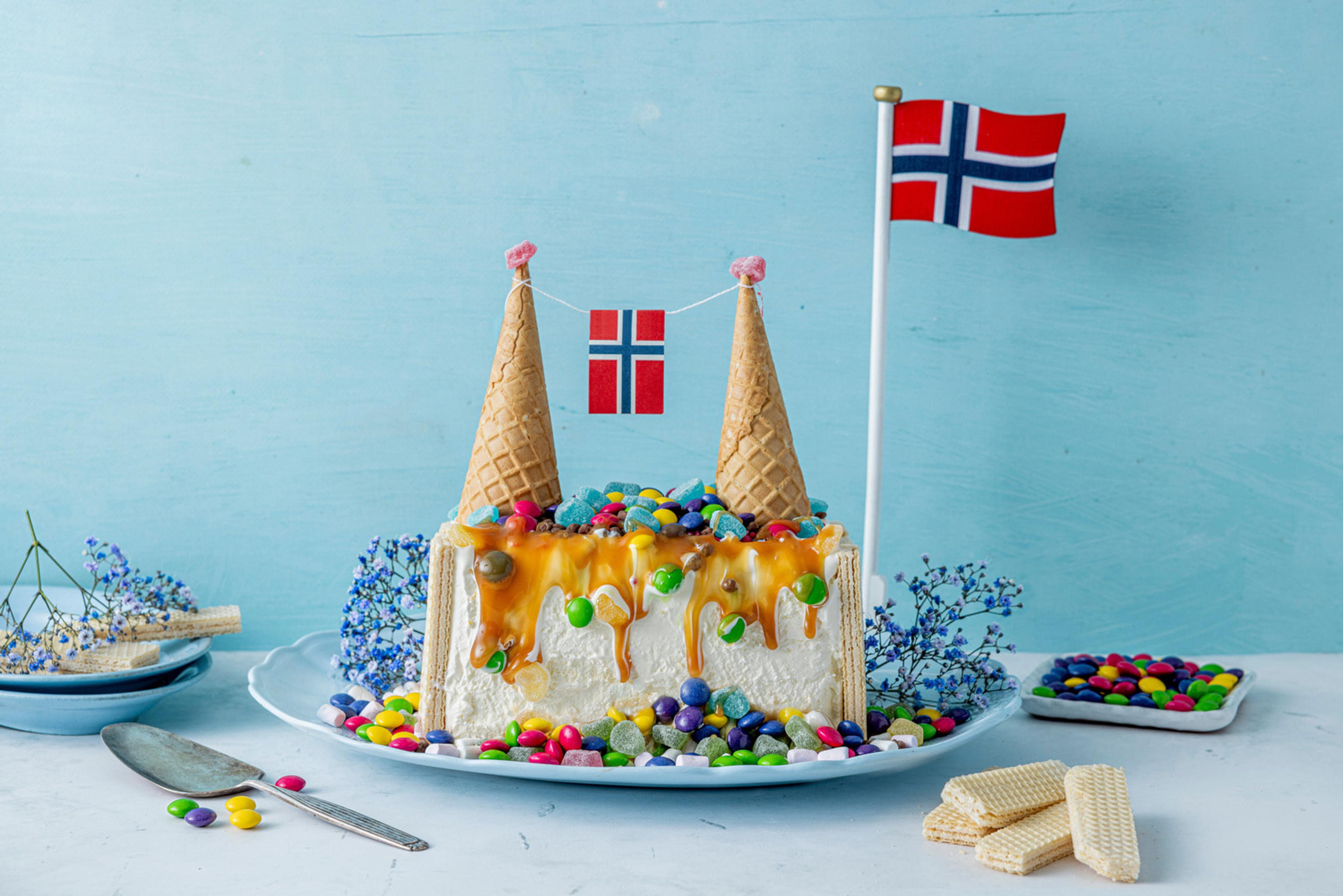 Iskake pyntet til festdager i mai, med tårn av iskjeks som holder det norske flagget. Isen er dekket i karamellsaus som renner langs sidene og godteri.