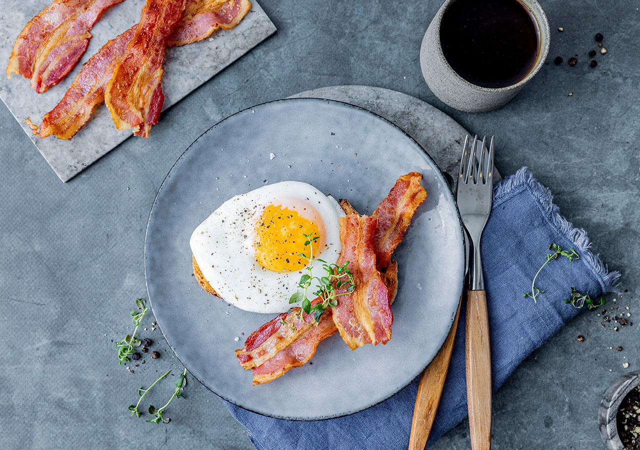 Stekt egg og bacon servert på brødskive med kaffe ved siden av.