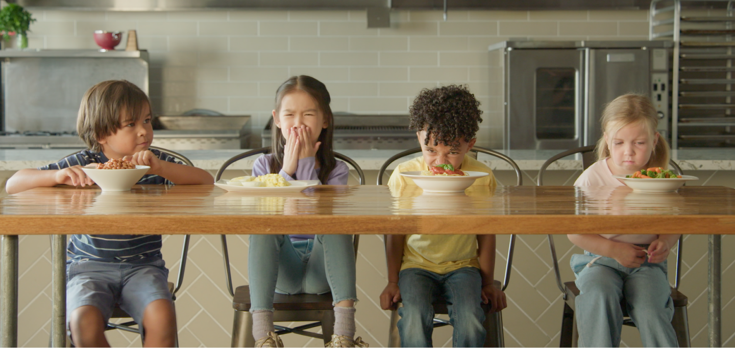 Four school age children sit in a kitchen filled with film lights and camera equipment 