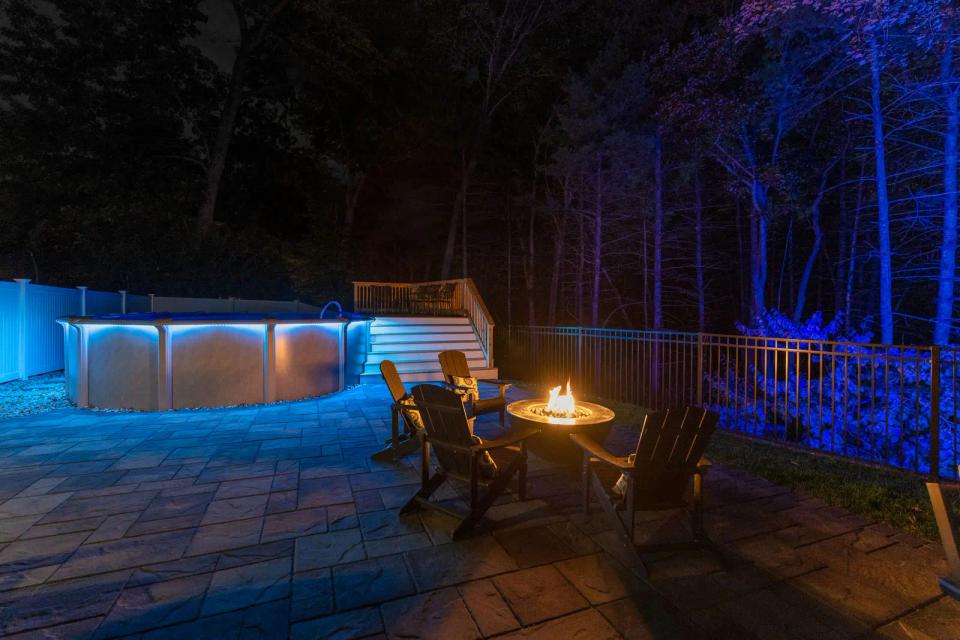 Above-ground pool and staircase illuminated in blue beside patio chairs and a glowing fire pit