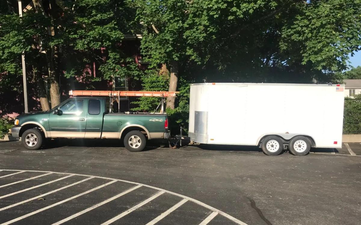Older green Ford F-150 with an orange ladder rack towing a white enclosed trailer