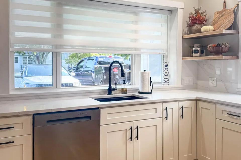 Black kitchen faucet beneath a window with white cabinetry and open corner shelves