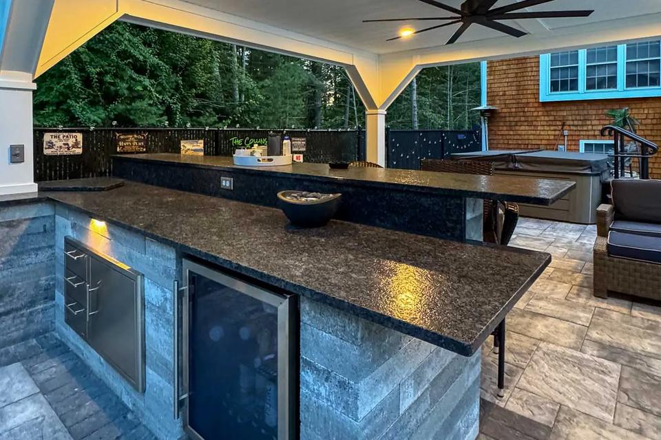 Dark bar countertop above a beverage fridge beneath the lit pavilion ceiling and black fan