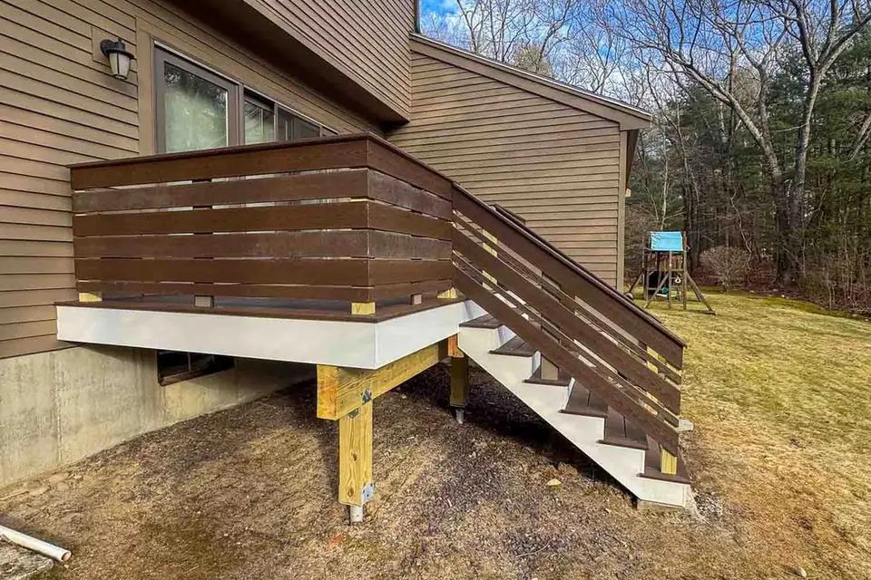 Elevated deck with dark horizontal railings and grey stairs beside a brown-sided home