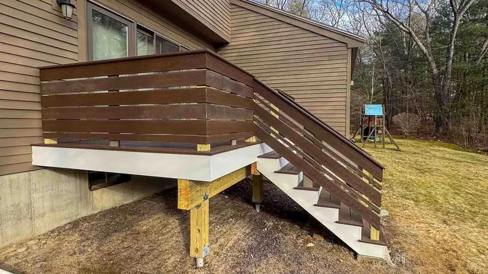 Elevated deck with dark horizontal railings and grey stairs beside a brown-sided home