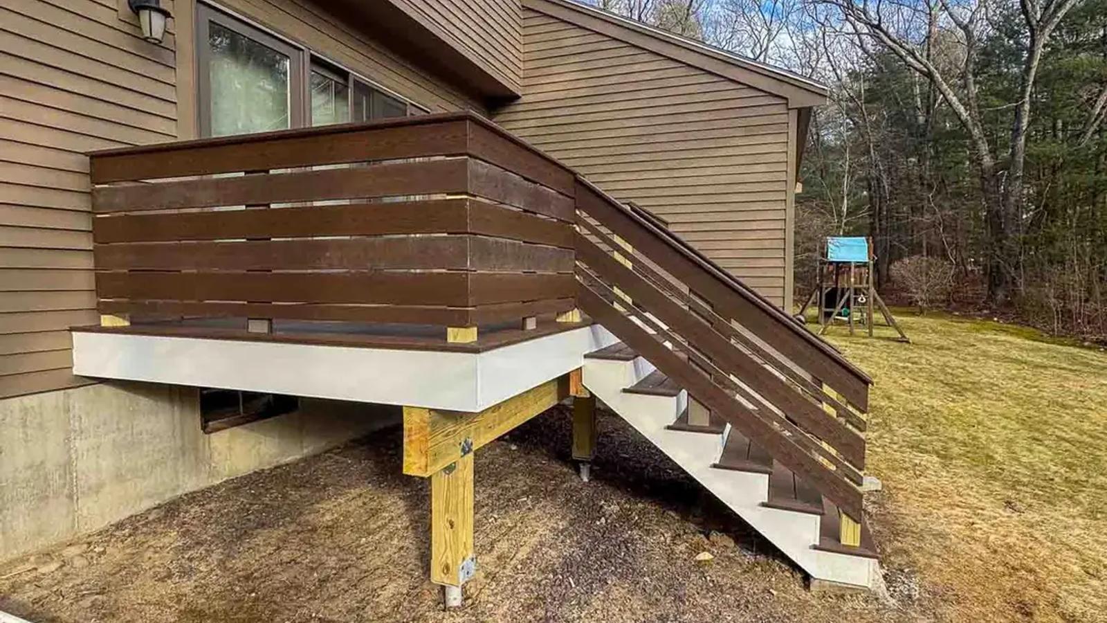 Elevated deck with dark horizontal railings and grey stairs beside a brown-sided home