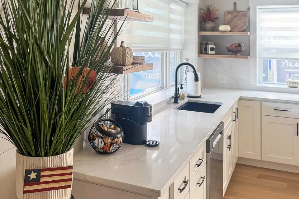 White countertop with a black sink, open corner shelves and a plant beside the window