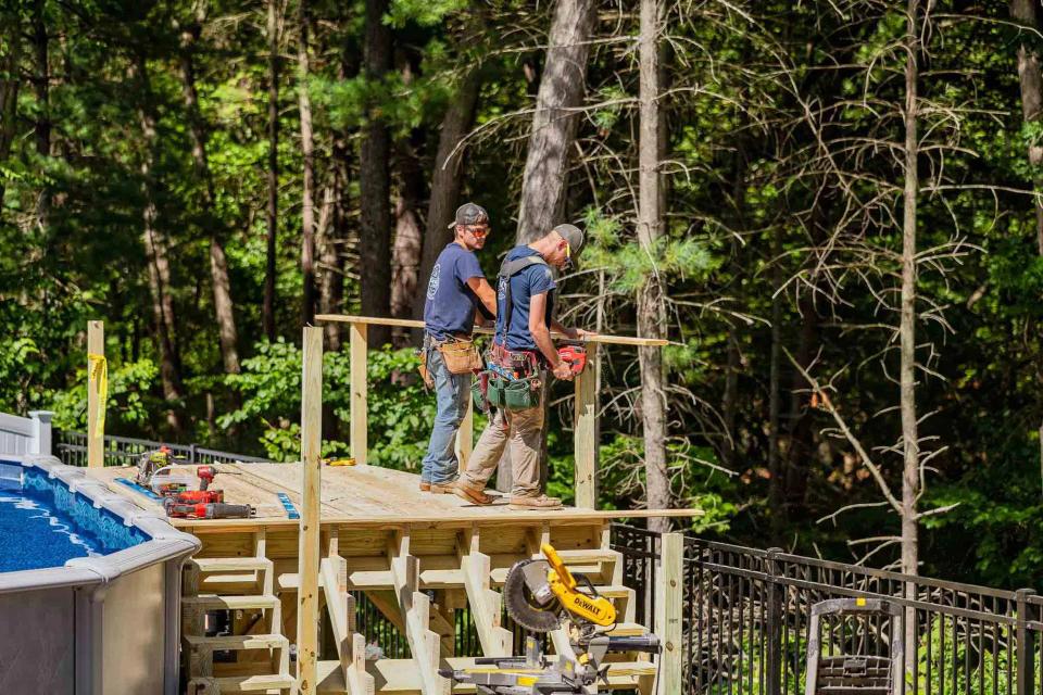 Two carpenters working on the wood deck frame beside an above-ground pool