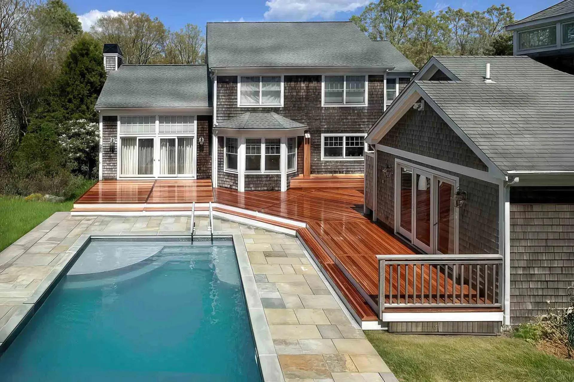 In-ground pool and stone patio beside a mahogany deck wrapping the shingled home