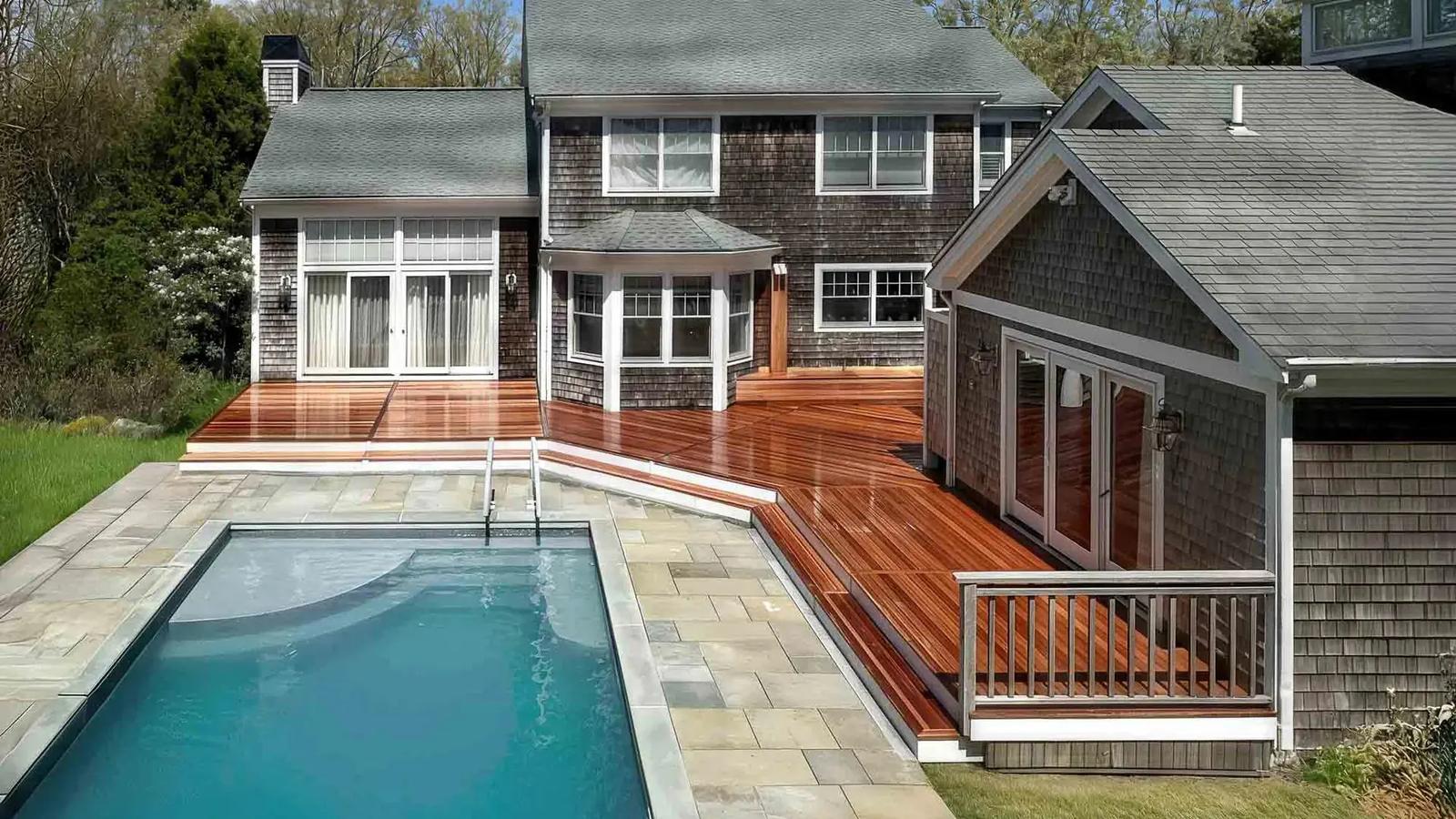 In-ground pool and stone patio beside a mahogany deck wrapping the shingled home