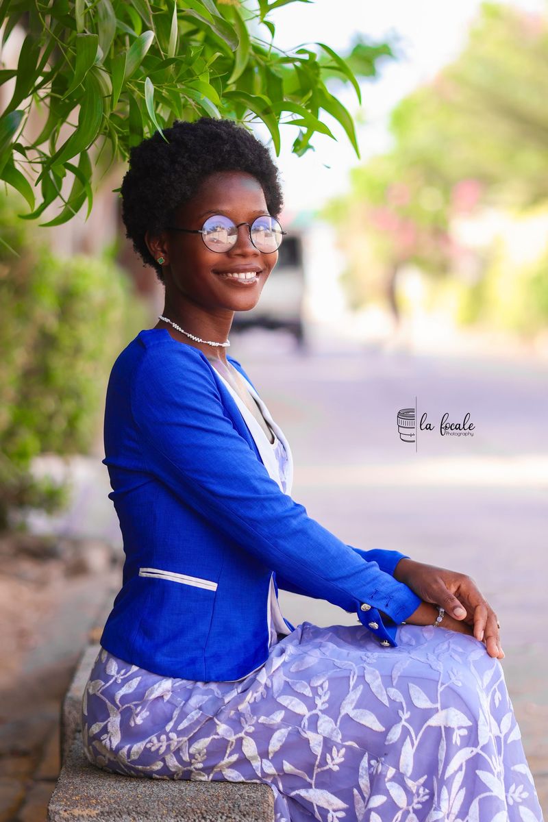 A young woman in a blue blazer and patterned skirt sits outdoors under green foliage, smiling