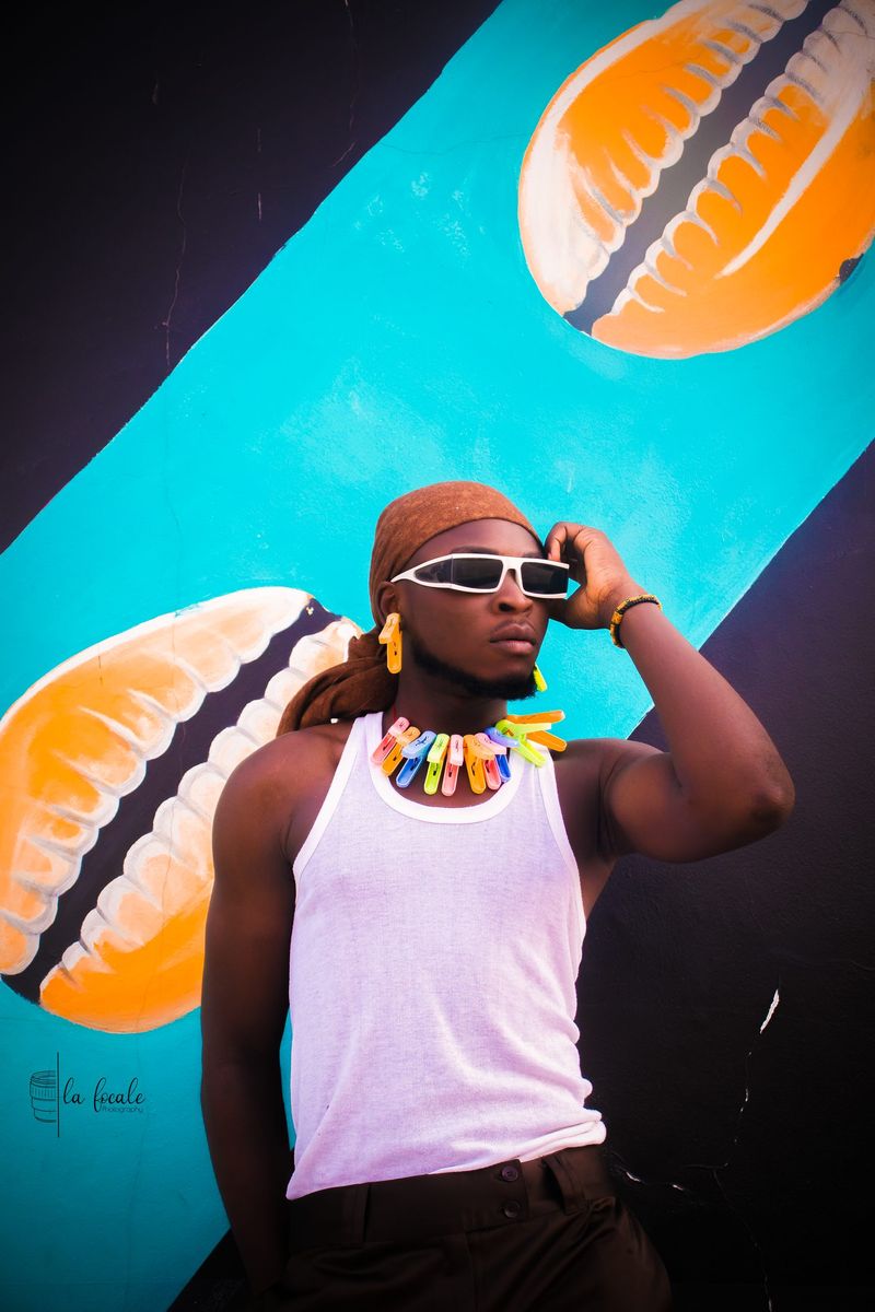 A young man in white sunglasses and a beaded necklace poses against a bright turquoise mural