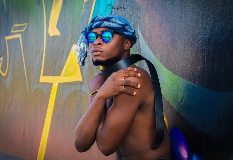 A young man in a blue turban and mirrored sunglasses poses with arms crossed against a graffiti wall