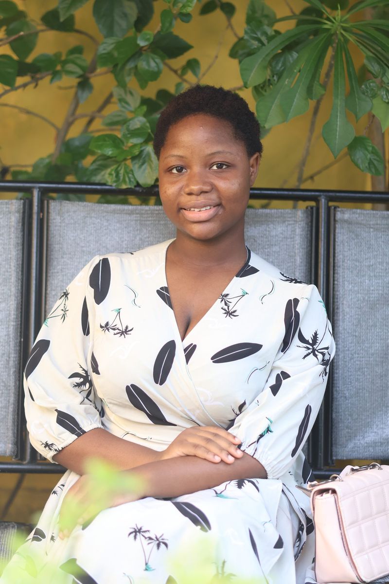 A smiling young woman in a leaf-print dress sits on a bench by a leafy green wall