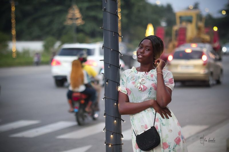 A young woman in a floral dress poses by a fairy-lit lamppost at dusk with city traffic behind