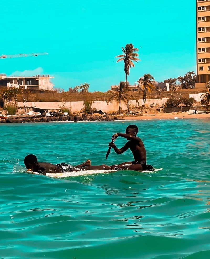 Two boys play on a surfboard in turquoise coastal water with a city skyline behind