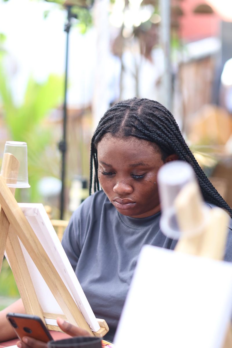 A young woman in a grey top checks her phone beside a small canvas easel