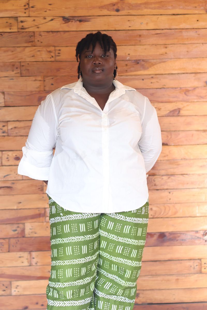 A guest in a white shirt and green patterned trousers stands against a wooden plank wall
