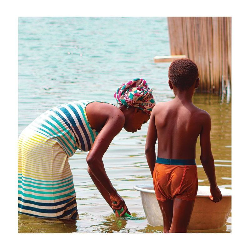 A woman in a striped dress and a boy stand in shallow water beside woven reeds, a basin nearby