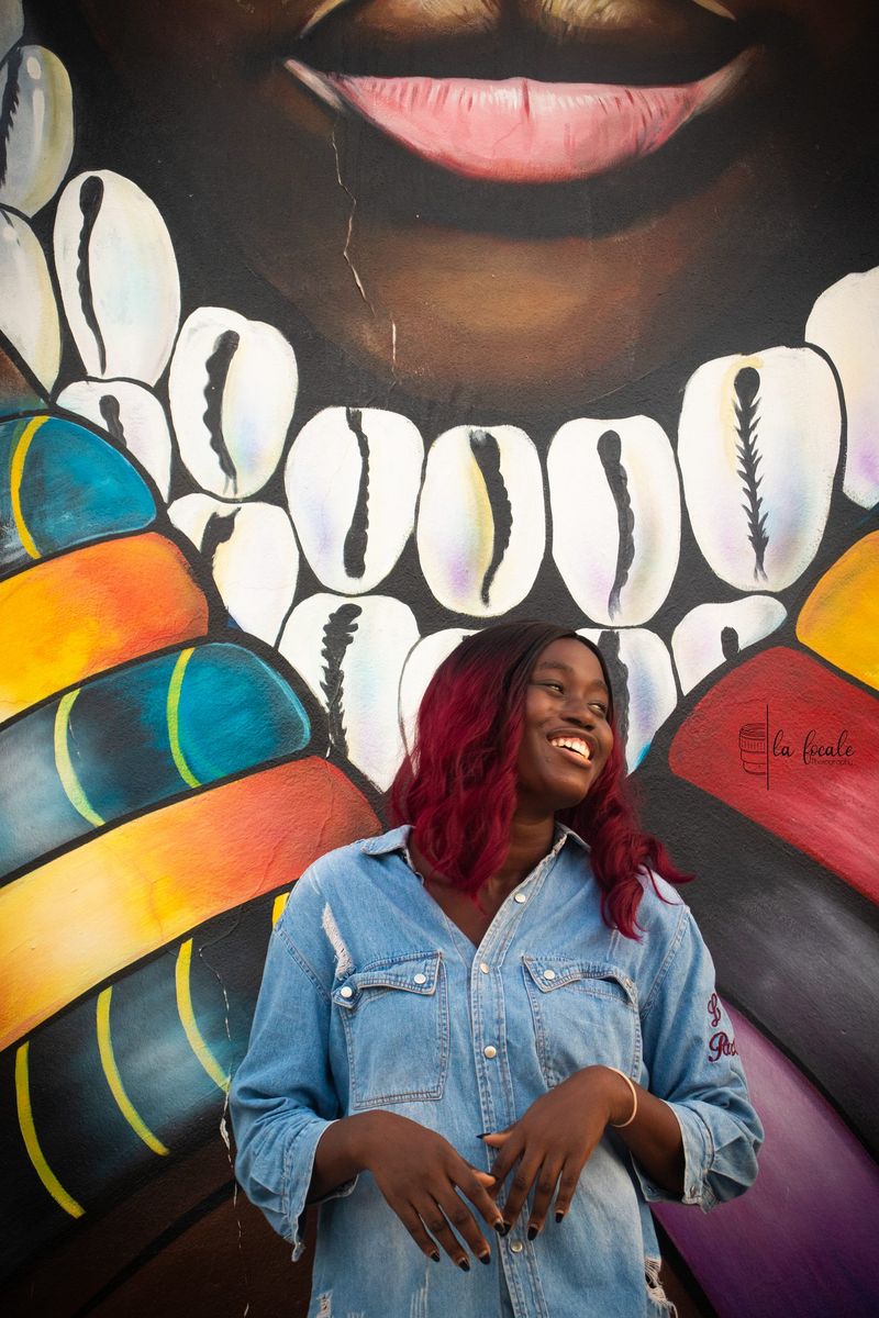 A smiling young woman in a denim shirt poses in front of a vibrant mural of cowrie shells