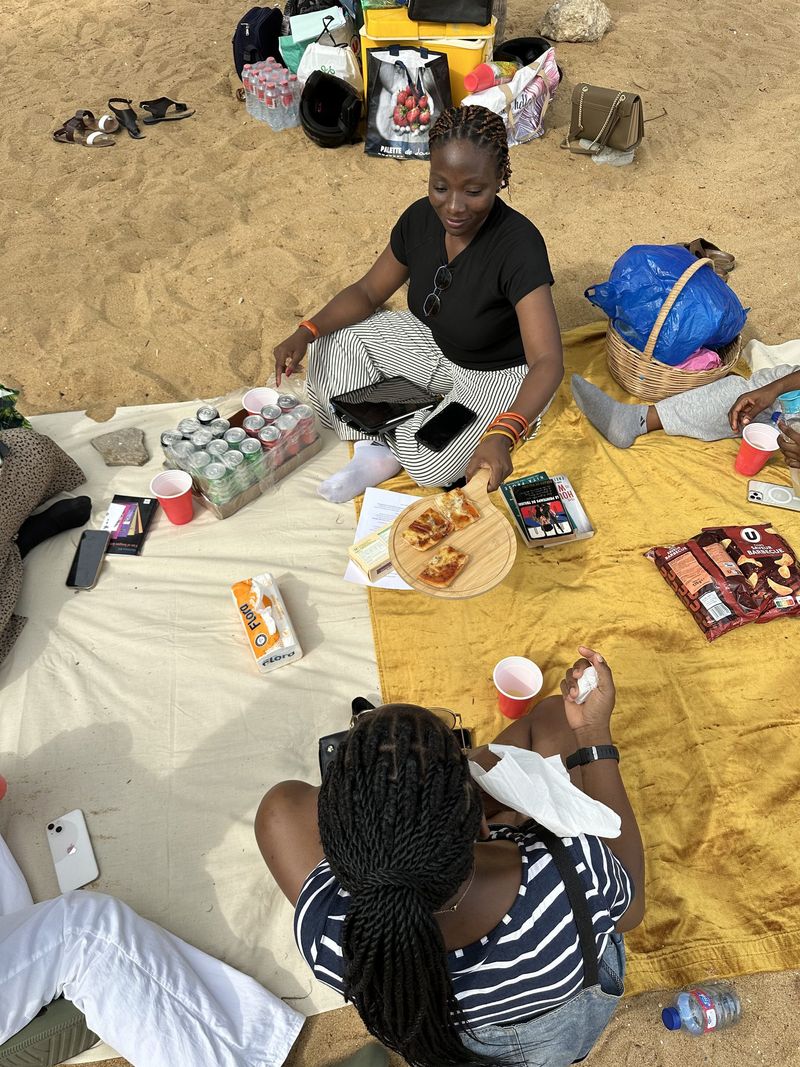Overhead view of two friends on a picnic blanket with food, drinks and books on the sand