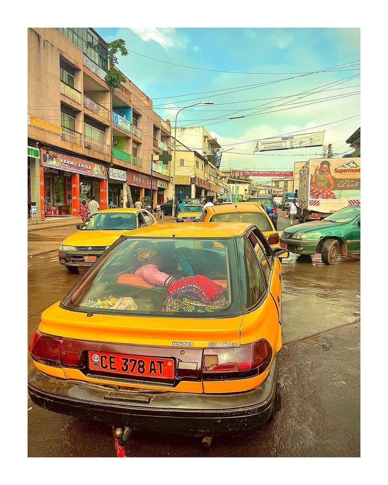 Yellow shared taxis in heavy traffic on a busy Central African city street lined with shopfronts and a billboard