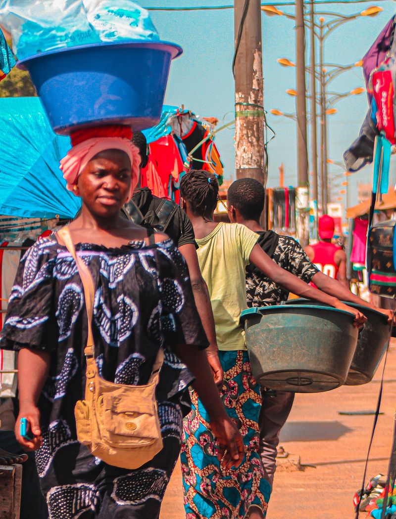 A woman balances a large blue basin on her head while walking through a busy market street