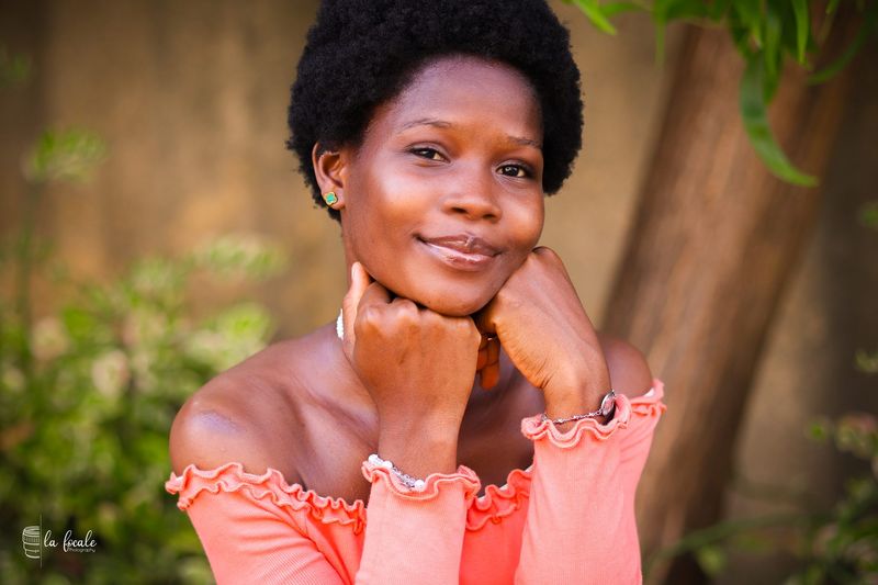 Portrait of a young woman in a coral off-shoulder top resting her chin on her hand, greenery behind