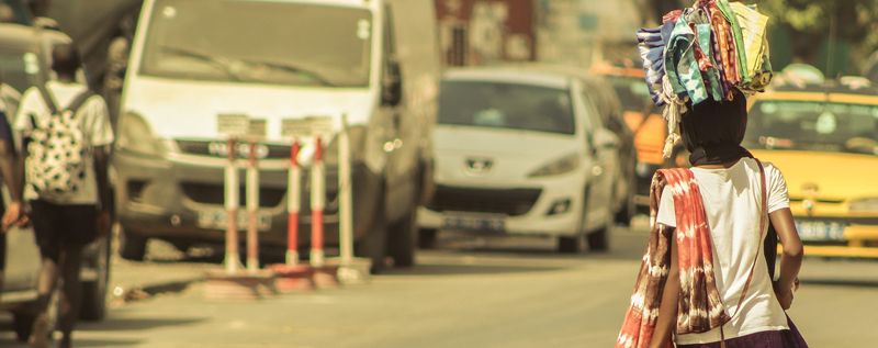 A street vendor carries a load on their head between cars in traffic