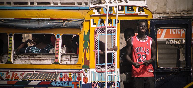 A young man in a red basketball jersey stands beside a brightly painted public minibus