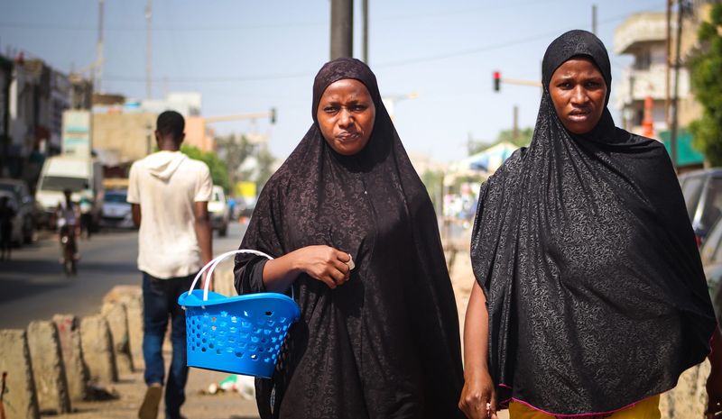Two women in dark robes walk along a busy street, one carrying a blue basket