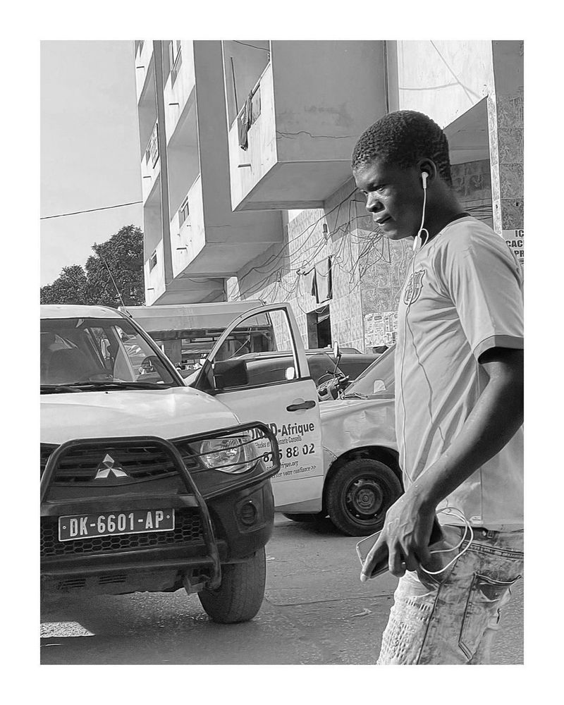 A young man with earphones stands beside a white pickup truck on a city street, black and white