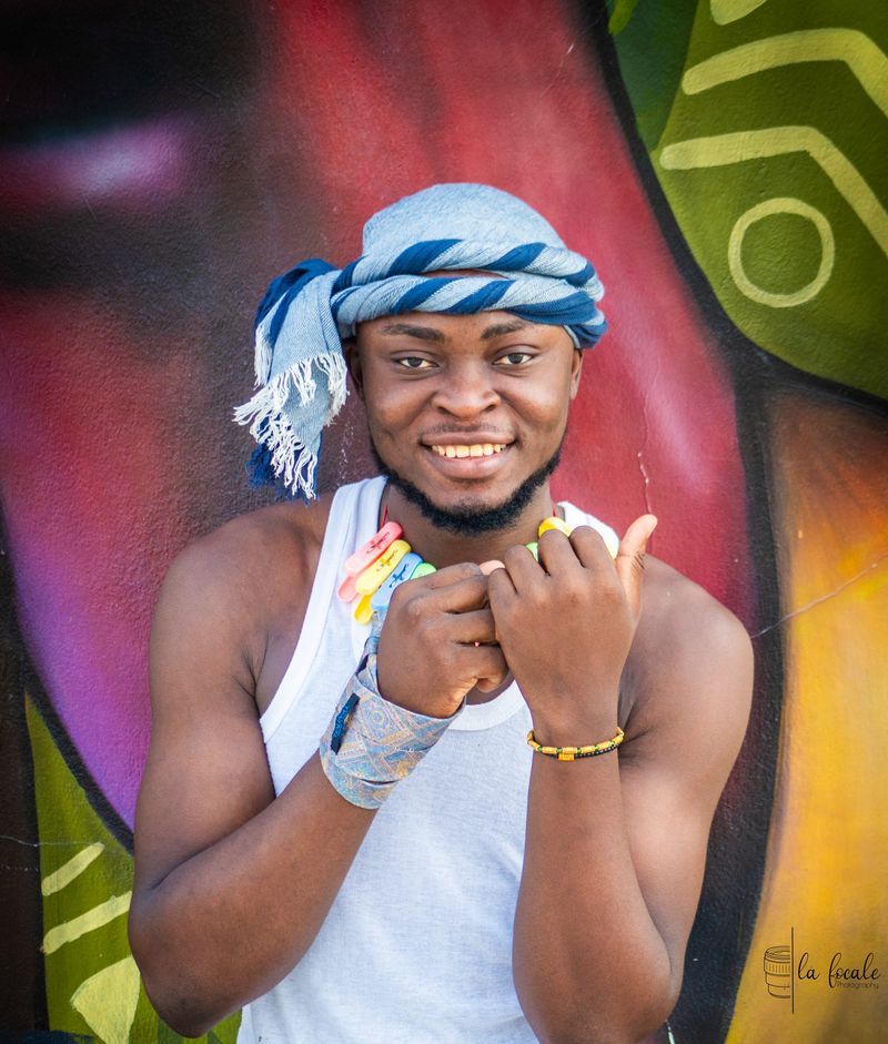 A smiling young man in a blue head-wrap and white tank top poses before a colourful mural