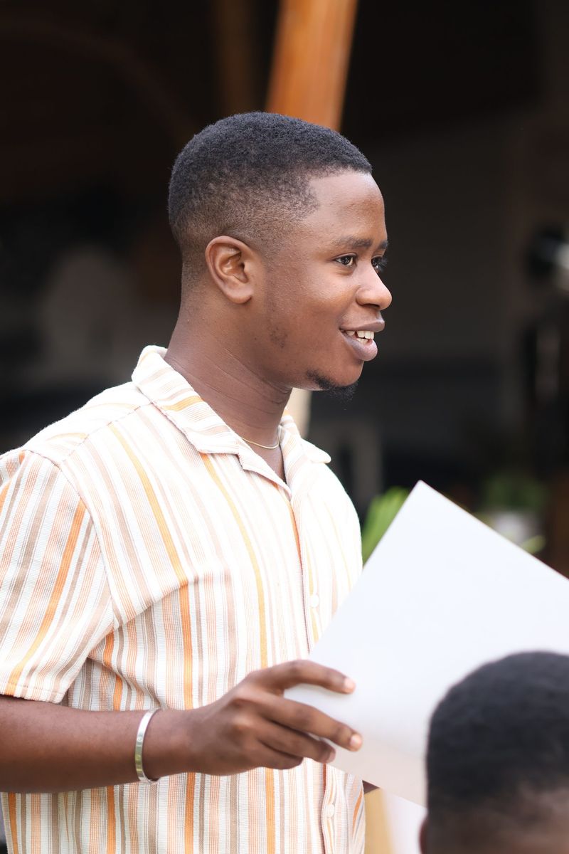 A young man in a striped shirt reads from a sheet of paper while speaking