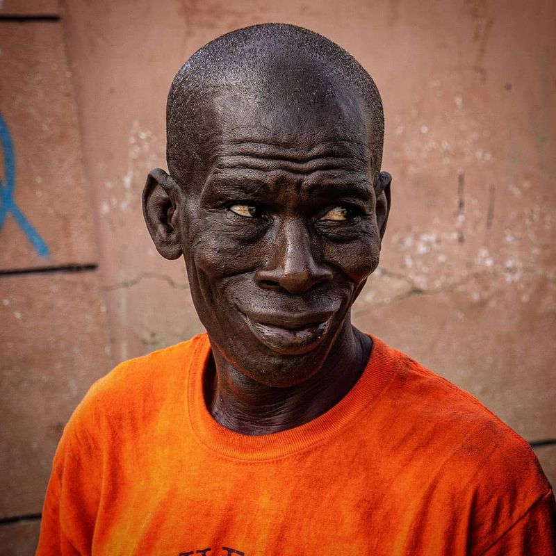 Close-up portrait of an older man in an orange t-shirt against a weathered wall, looking into the camera