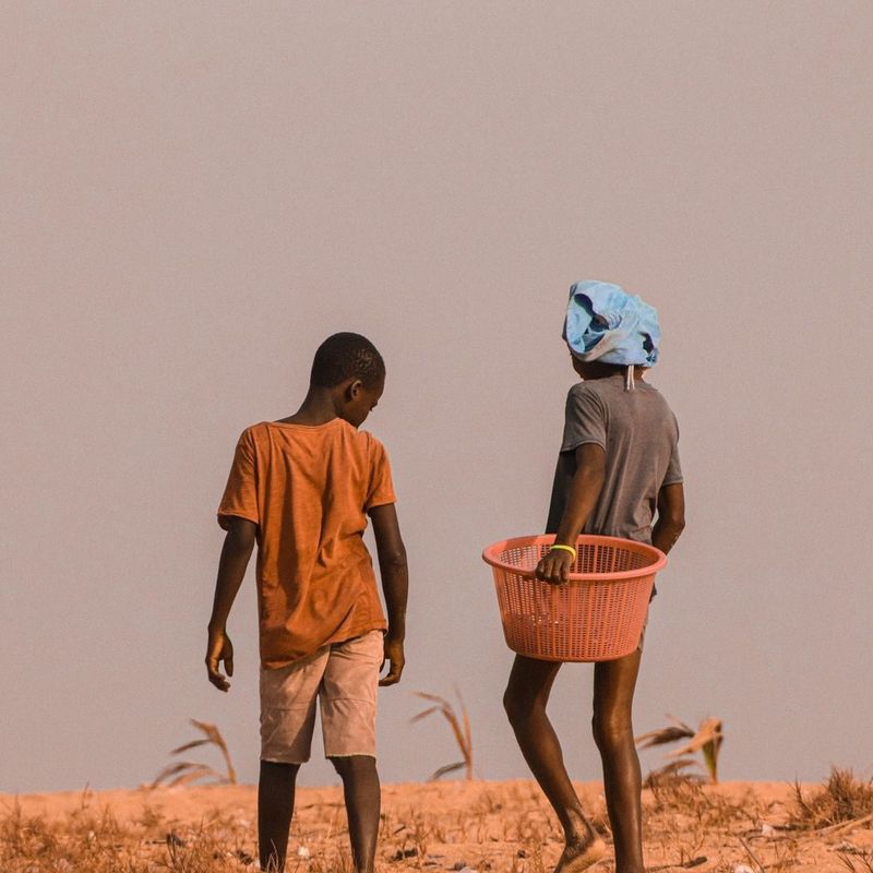 Two children walk along a path, a boy in an orange t-shirt and a girl carrying an orange basket, warm-toned