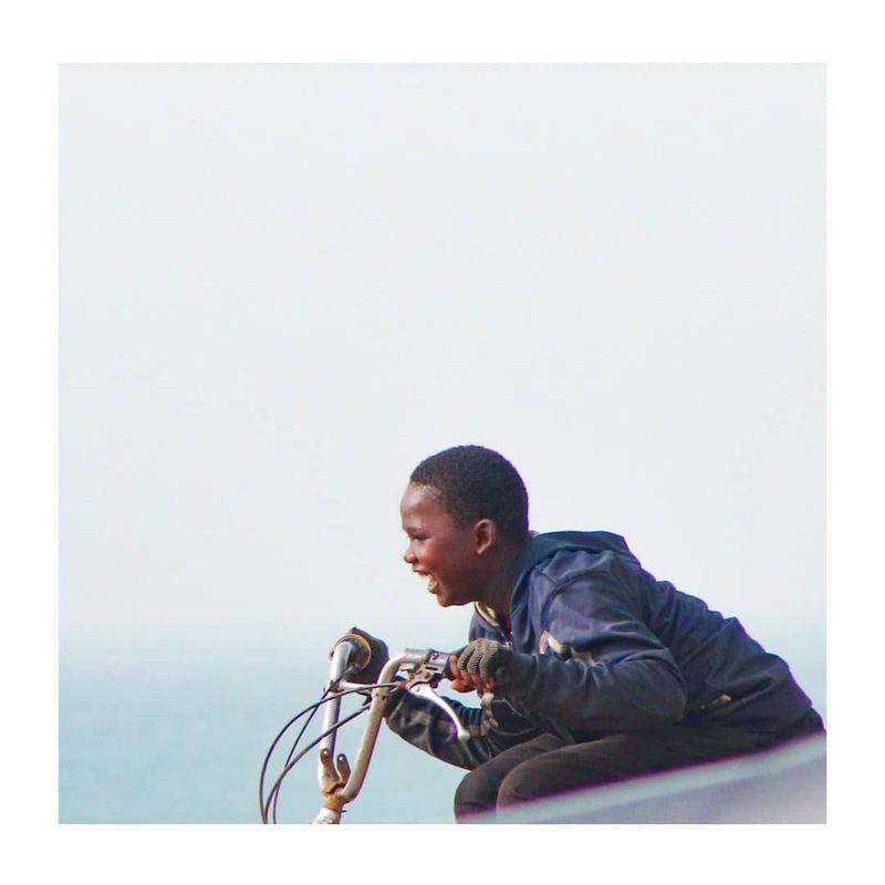 A smiling boy rides a bicycle along the seafront against a bright hazy sky