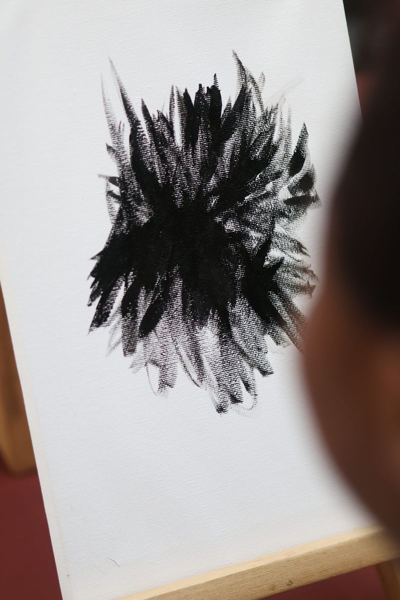 A canvas on an easel shows a bold black brushwork painting resembling a sunflower