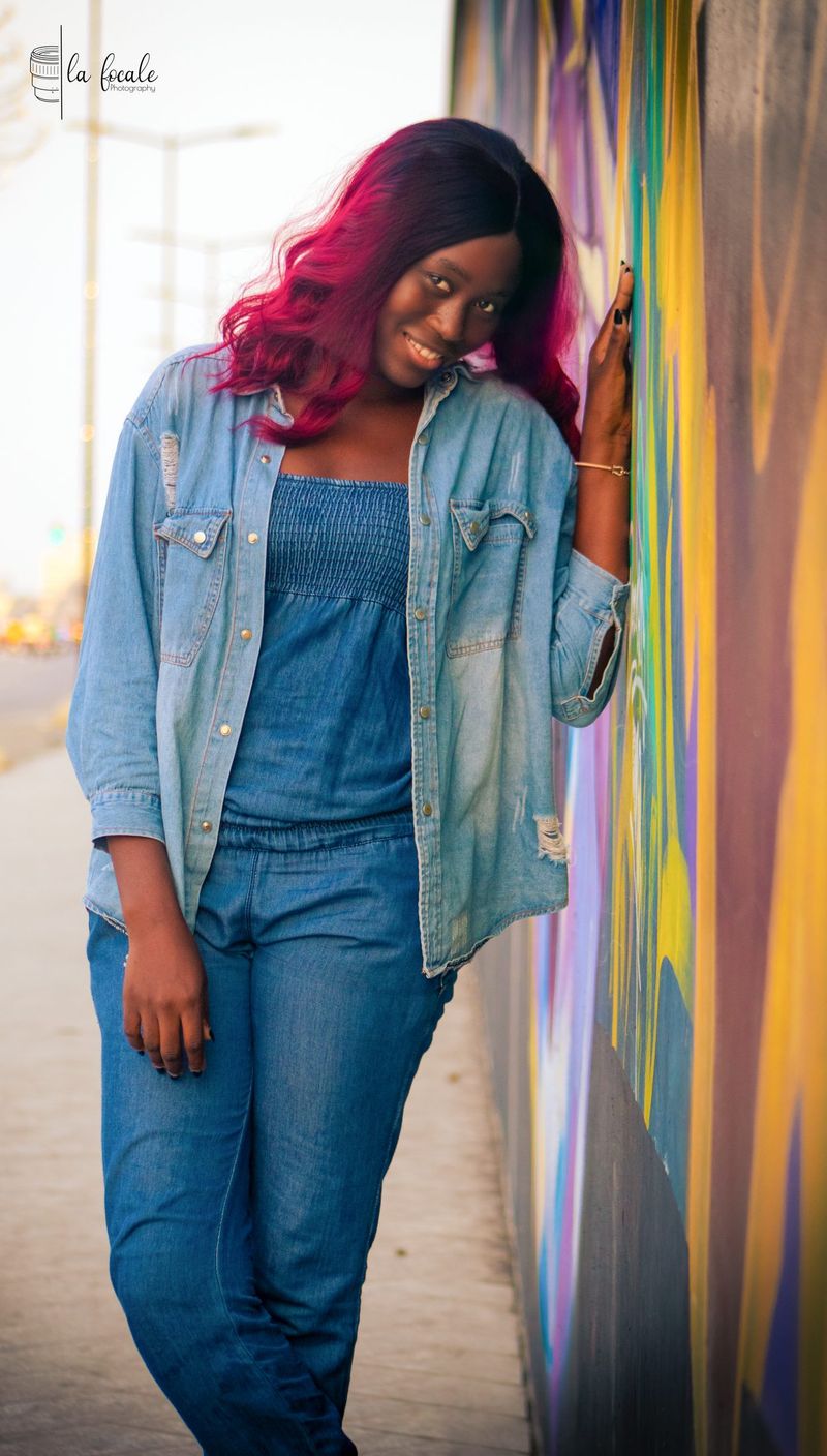 A young woman in a denim jacket leans against a colourful graffiti wall, La Focale portrait