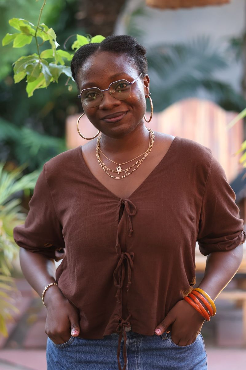 A young woman in a brown top and glasses smiles with her hands in her pockets in a garden setting