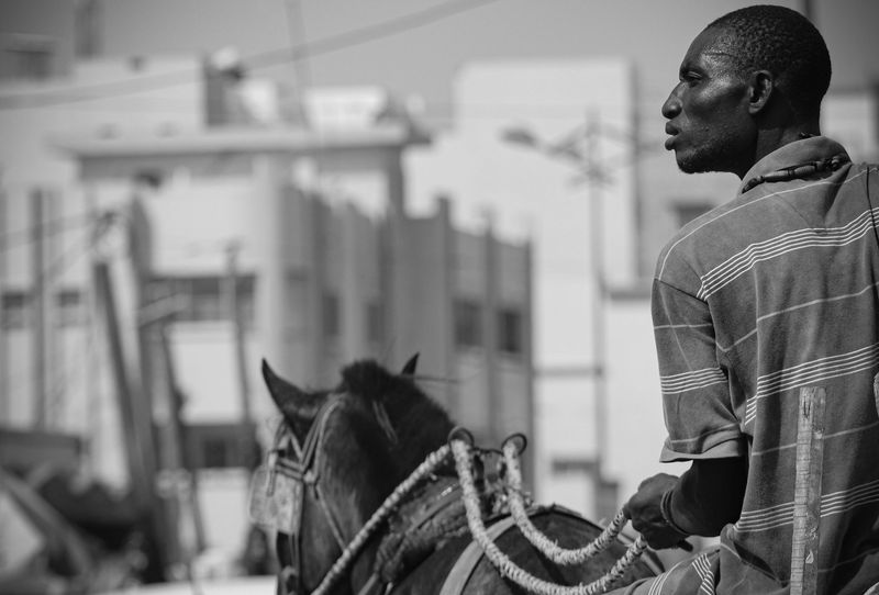 A man rides a horse through a city street, seen from behind, black and white