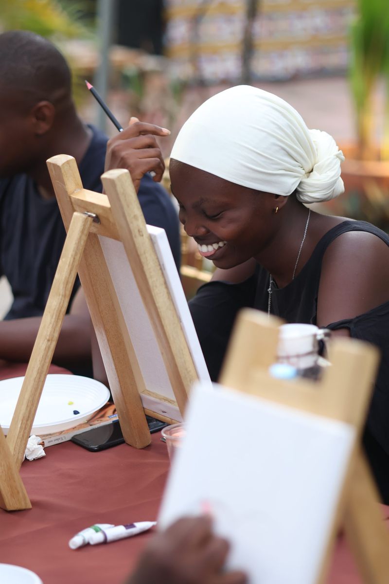A smiling woman in a white head-wrap paints at an easel among other guests