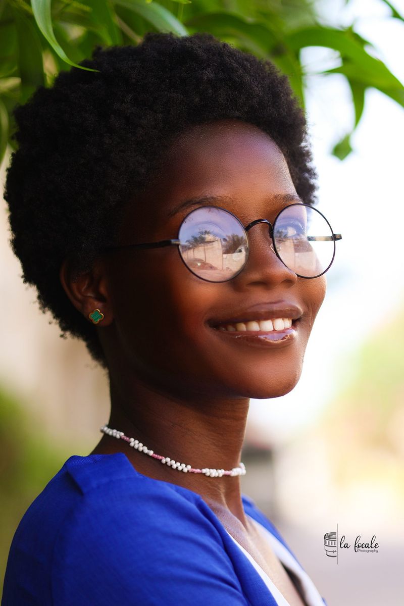 Portrait of a smiling young woman with round glasses and a blue top, greenery behind, La Focale photoshoot