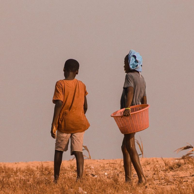 Two children walk across a dry field, one carrying an orange basket, warm-toned
